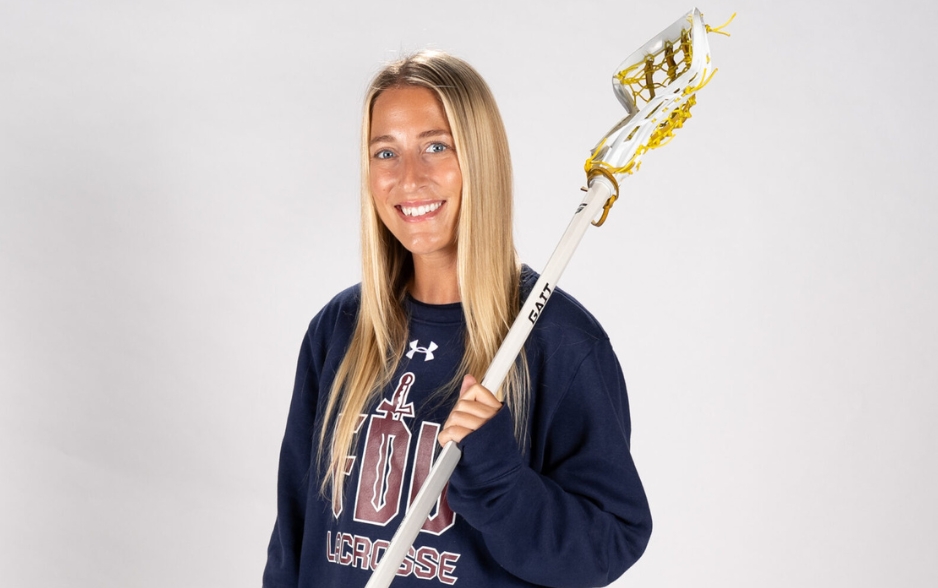 a woman stands in front of a blank backdrop and smiles at the camera. she holds a lacrosse stick and wears a FDU Lacrosse sweatshirt.