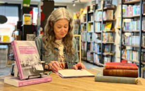 a woman sits at a table and signs a book. another copy of the book is displayed next to her.