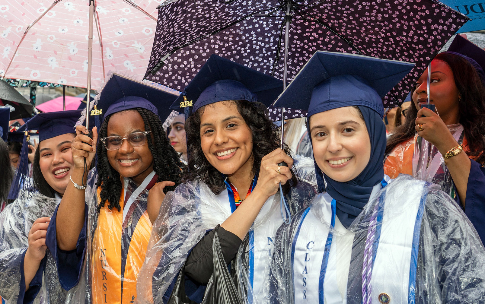 Four female graduates pose for a photo in the rain with their umbrellas.