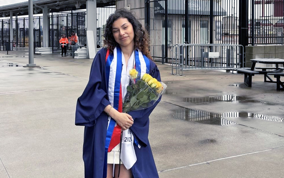 A young woman in a graduation robe poses for a photo outside of a stadium.