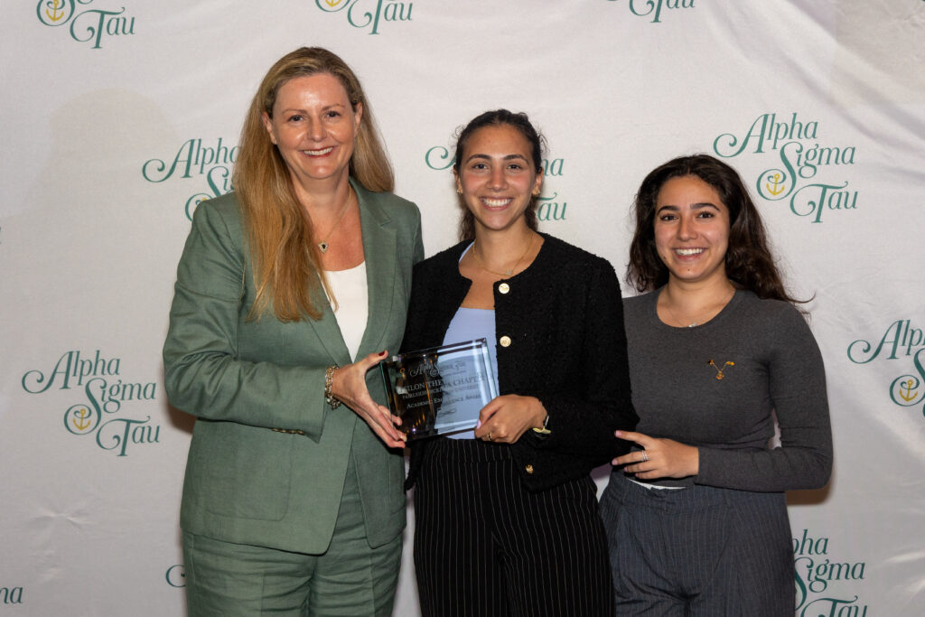 two students and a woman stand and smile for the camera. they are dressed in sorority badge attire. the woman is presenting the students with an award. the backdrop reads "Alpha Sigma Tau."