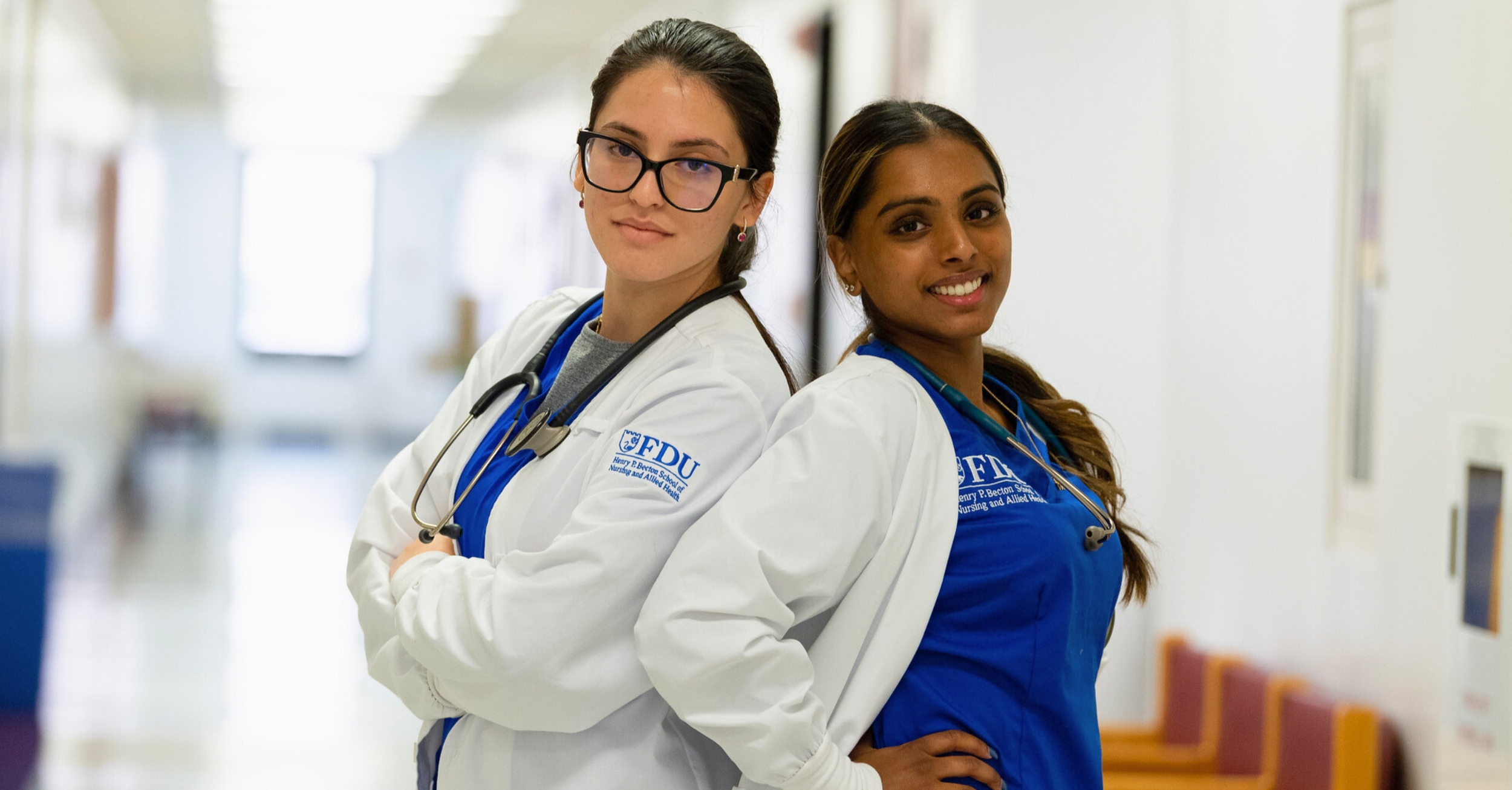 Two nursing students in white lab coats stand back-to-back in a bright hallway, smiling confidently. They wear stethoscopes and uniforms with the FDU School of Nursing and Allied Health logo.