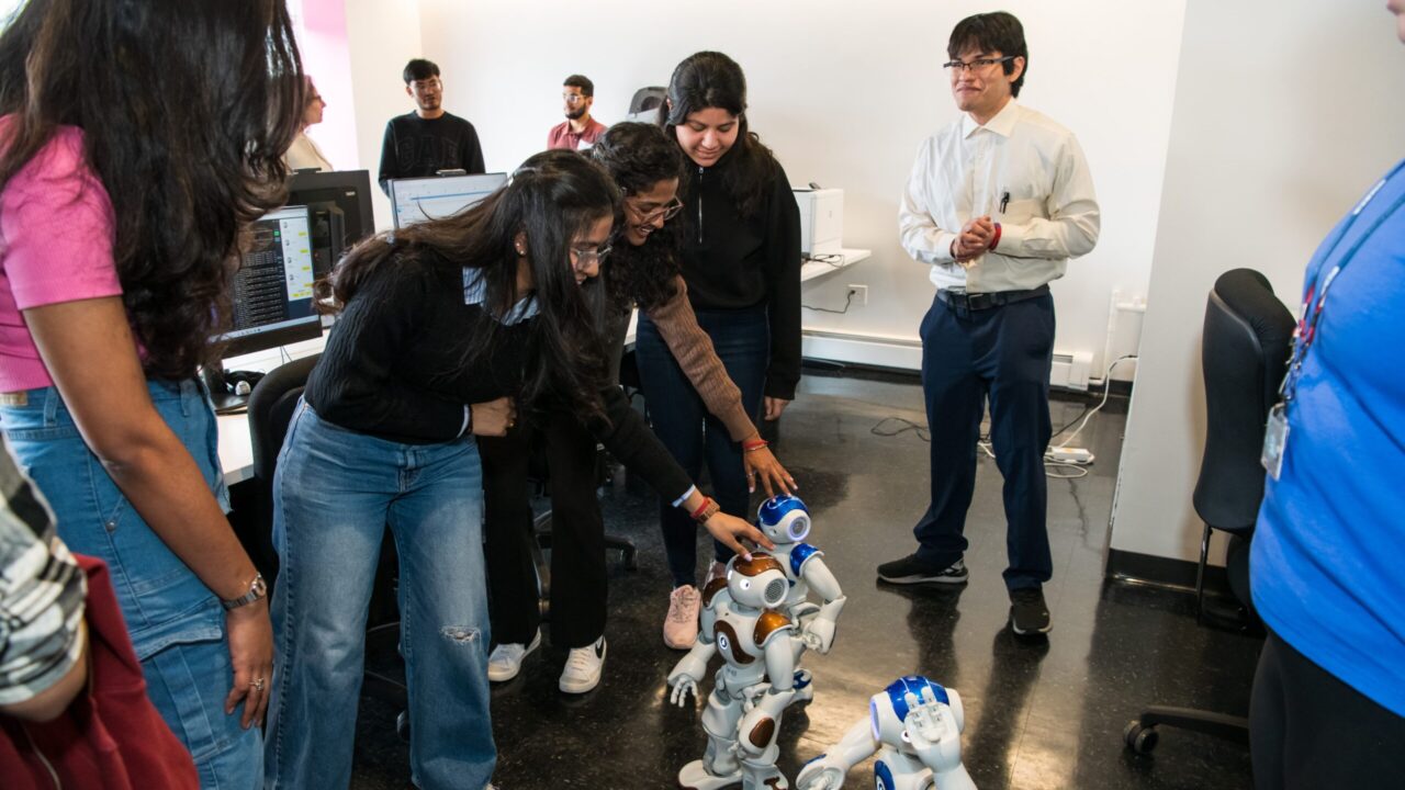 several students stand in a circle. they bend down to touch doll-sized robots.
