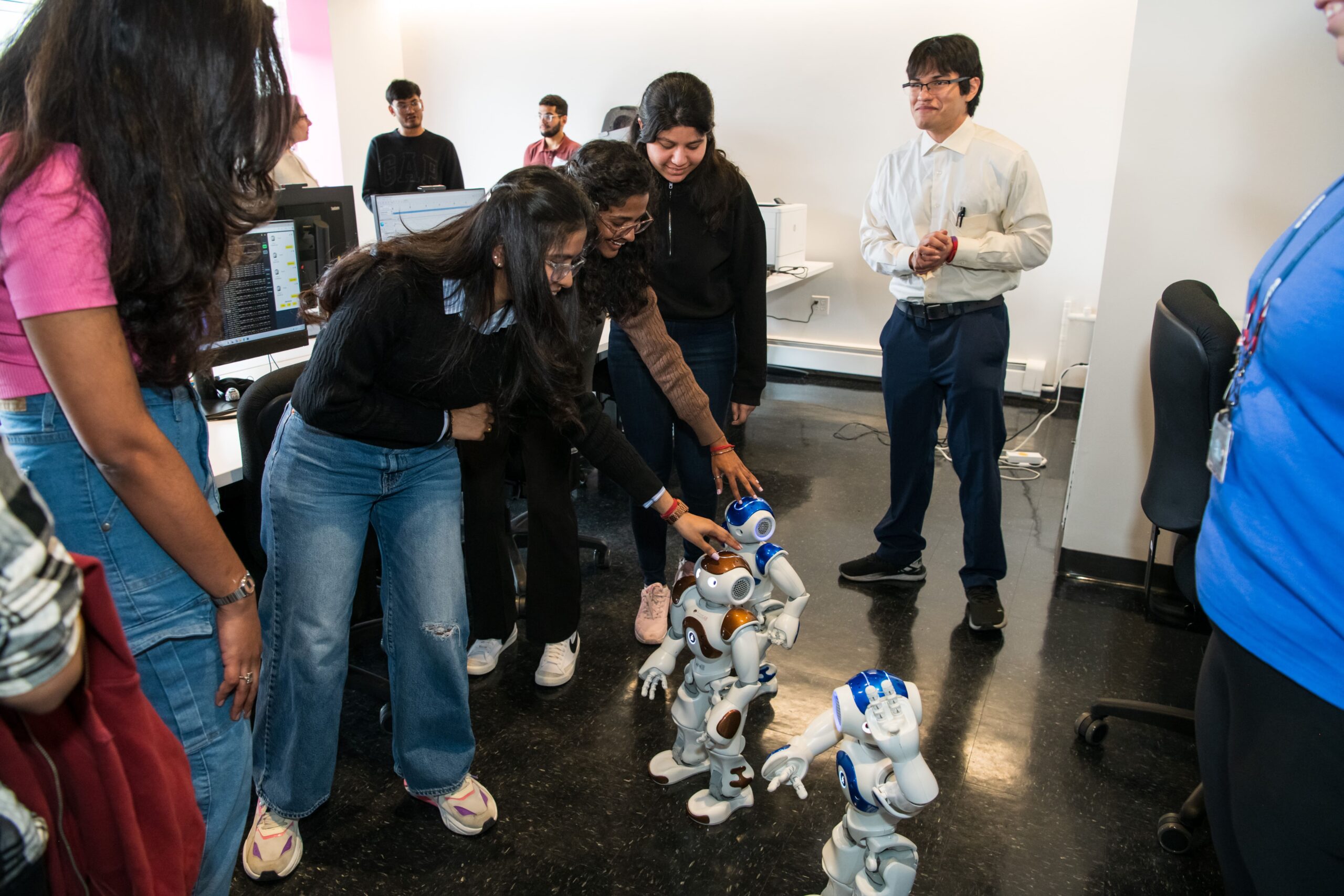 several students stand in a circle. they bend down to touch doll-sized robots.