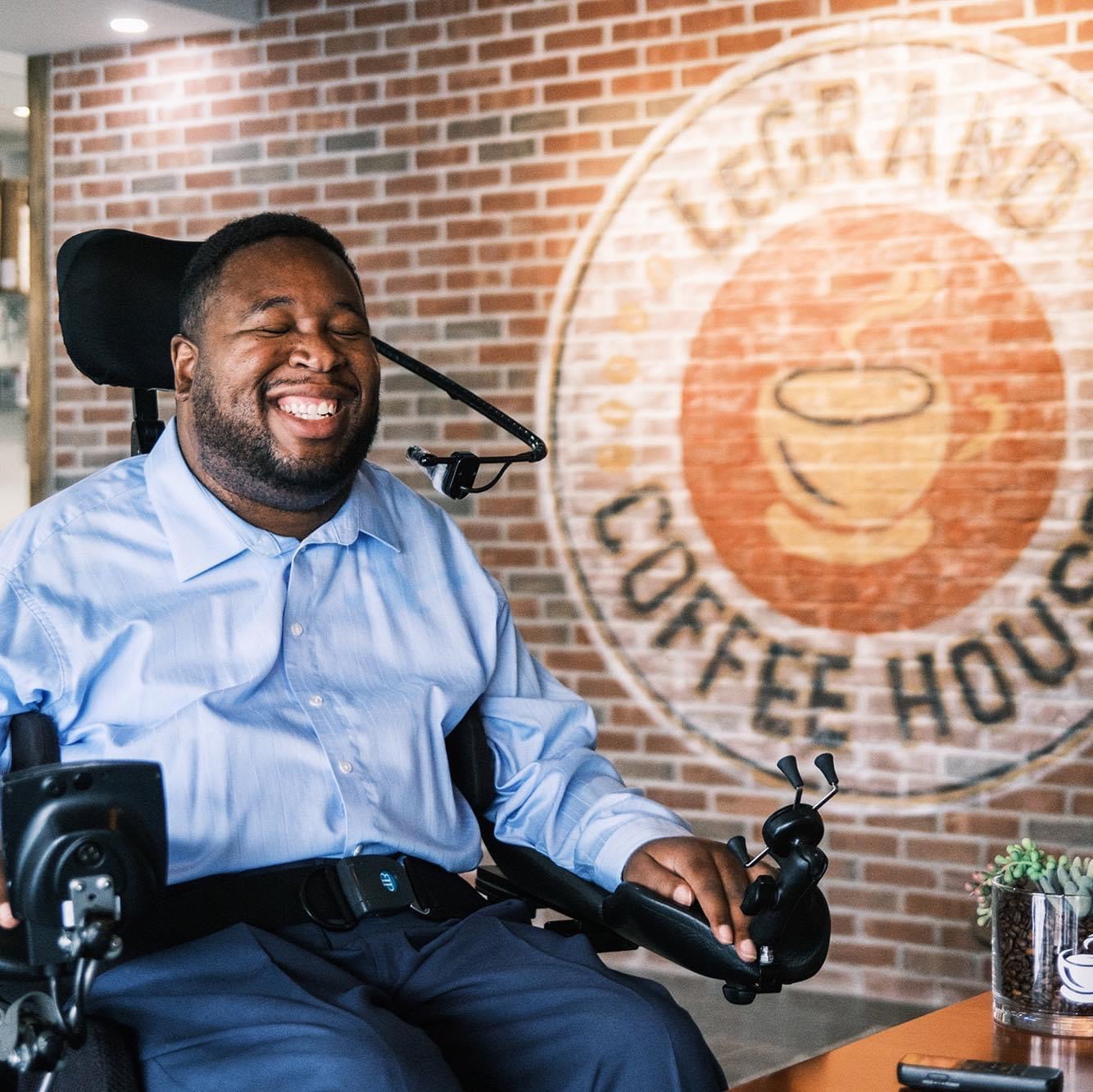 a man smiles at the camera. he sits in a wheelchair. there is a LeGrand Coffee House logo on the wall behind him.