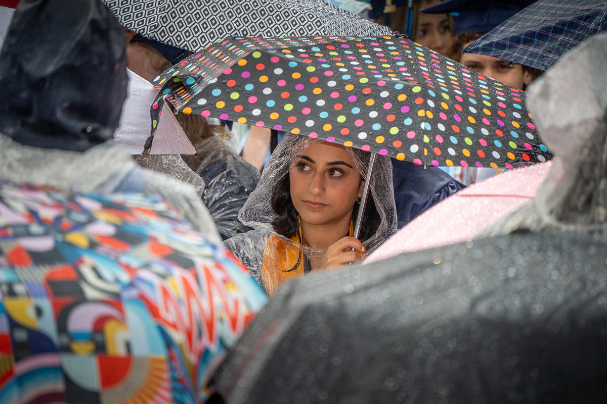 A young woman peeks out from under an umbrella, poncho and graduation cap.