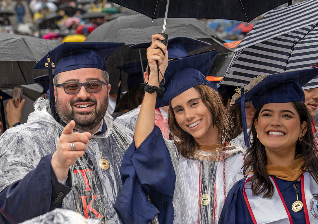 Three students in caps and gowns, with umbrellas and ponchos smile for a photo.