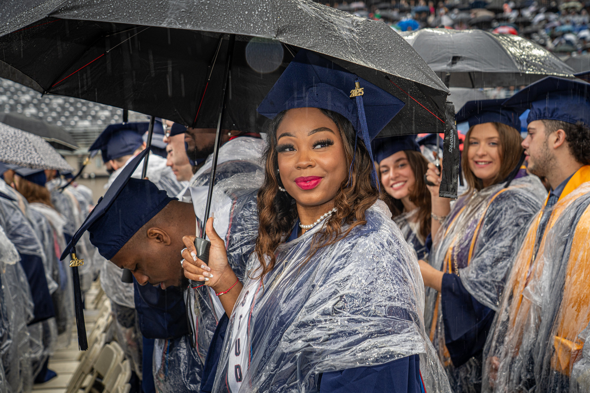 a graduate poses for the camera. she wears a rain poncho over her regalia and holds an umbrella.