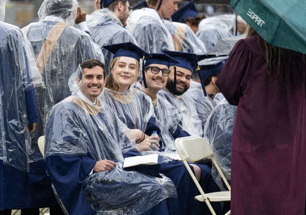 A group of students wearing ponchos with their caps and gowns smile.