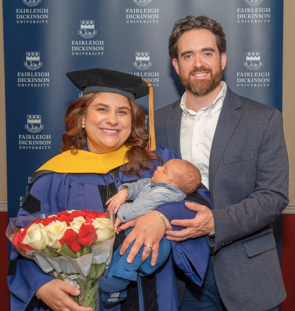 a family smiles for the camera. the mother wears doctoral robes as she holds roses and her baby.