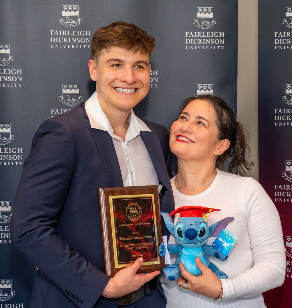 a student and his mother pose for the camera. the student holds an award while the mother holds a graduate Stitch plush toy. the mother smiles up at the student.