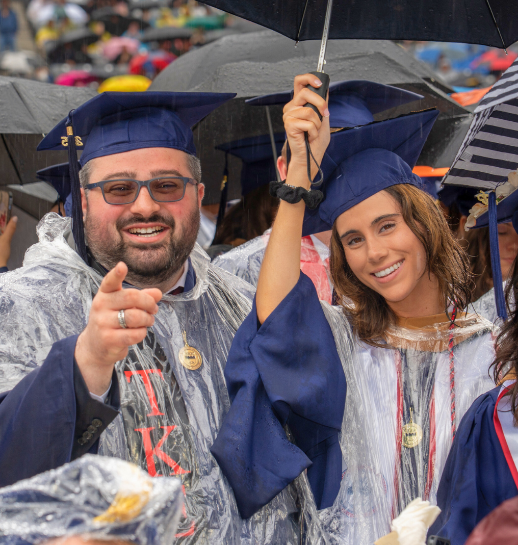 two students in caps and gowns, with umbrellas and ponchos smile for a photo.