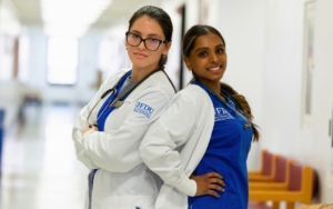 two students stand back to back. they both wear nurse uniforms and lab coats. one student crosses her arms and the other student has her hands on her hips.