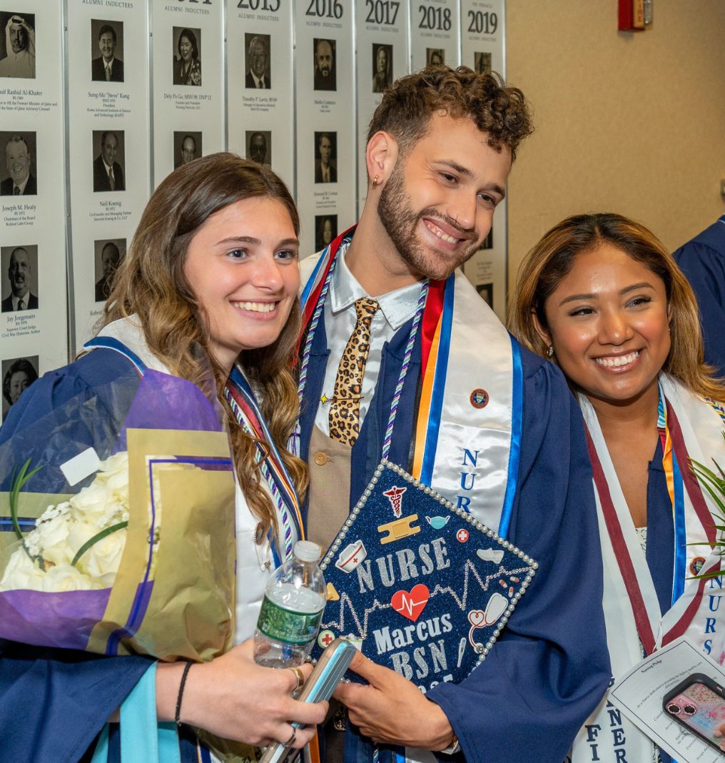 three graduating students smile for a camera out of frame. one holds flowers and another holds a grad cap that reads 