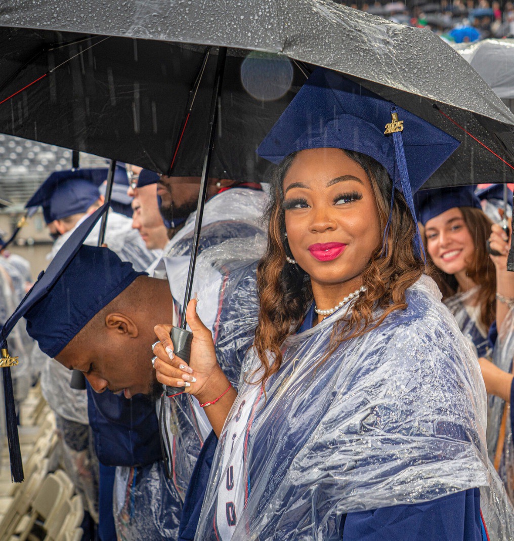 a graduate poses for the camera. she wears a rain poncho over her regalia and holds an umbrella.