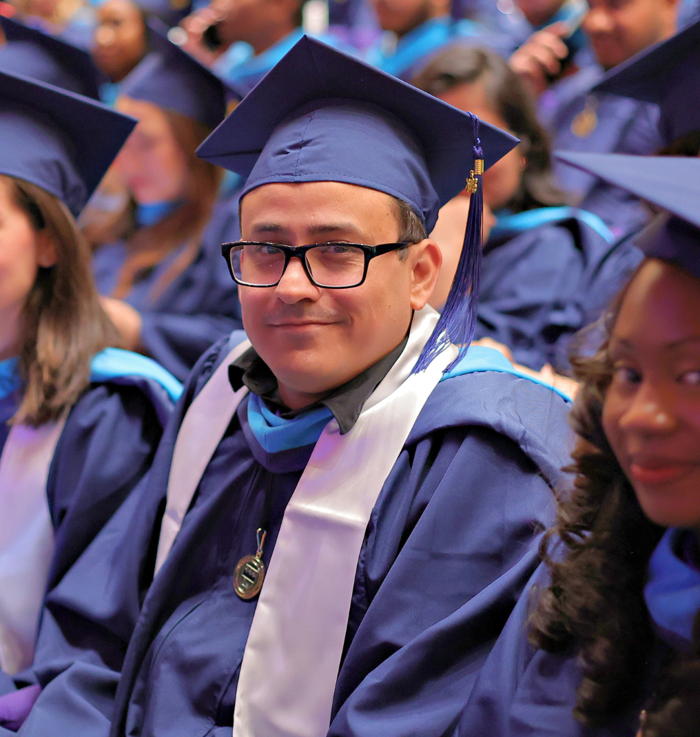 a graduate smiles at the camera. he wears glasses and regalia.