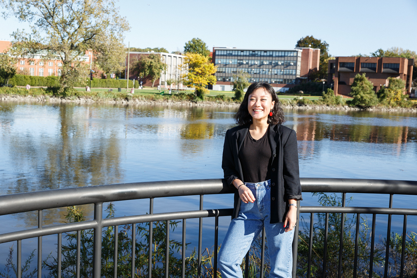 A student posing infront of the campus and a lake while leaning on a bridge.