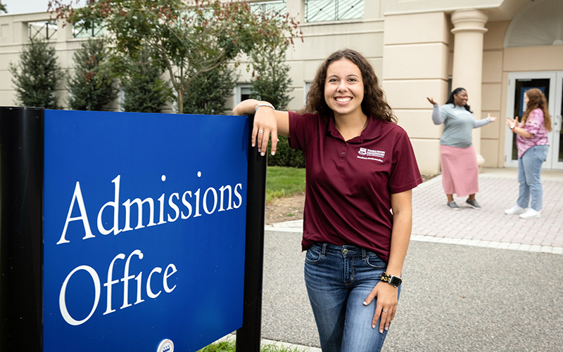 A woman drapes her arm over a sign for the Admissions Office.