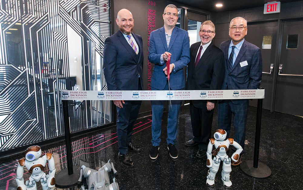 FDU President Michael Avaltroni and others cut a ribbon. they are all dressed in suits. they stand in front of a modern technology lab, surrounded by doll-sized robots.