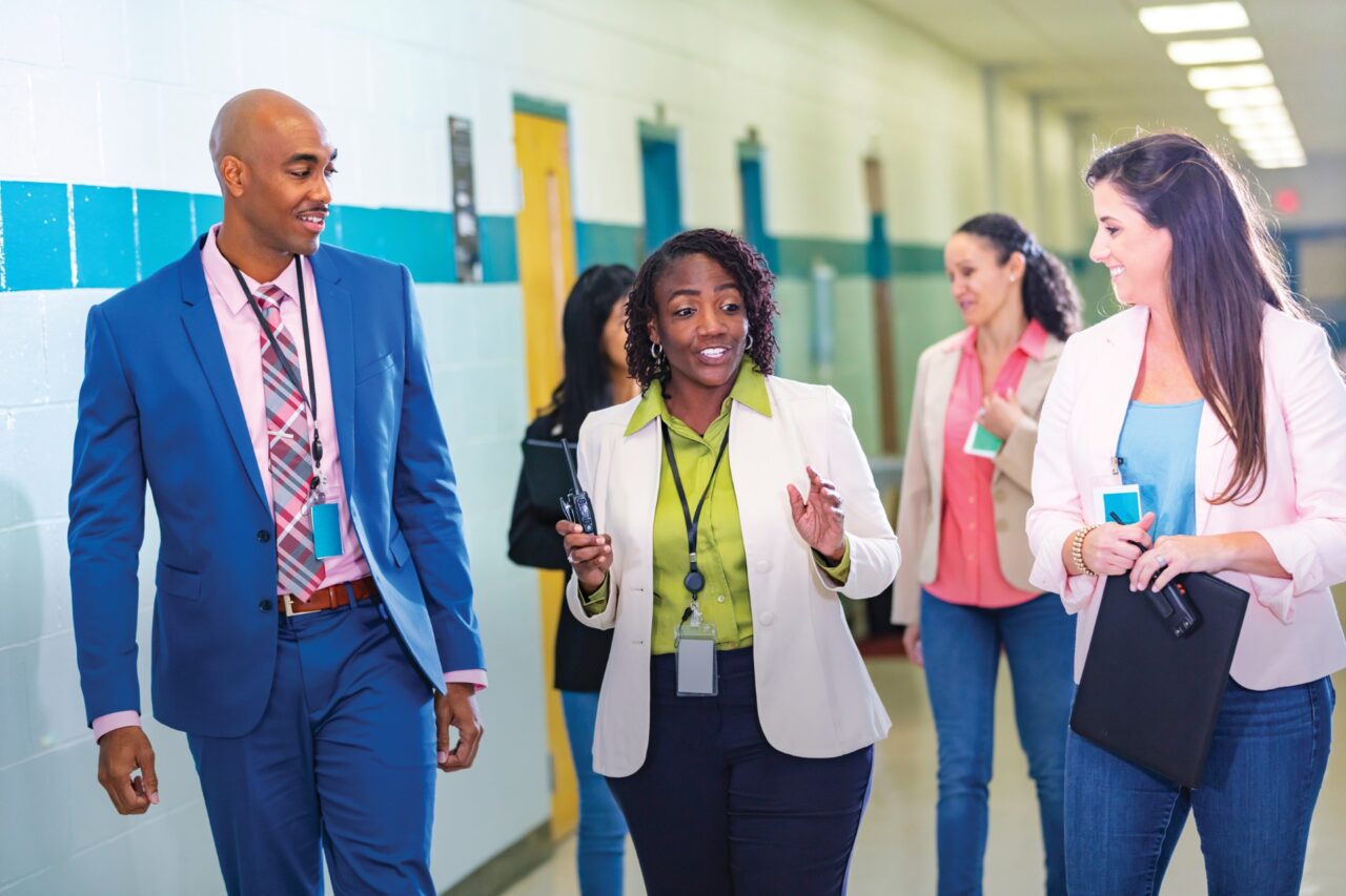 Four professionals in business casual attire walk down a brightly lit hallway, smiling and engaged in conversation. One holds a walkie-talkie, while another carries a folder. The setting appears to be an educational or office environment.