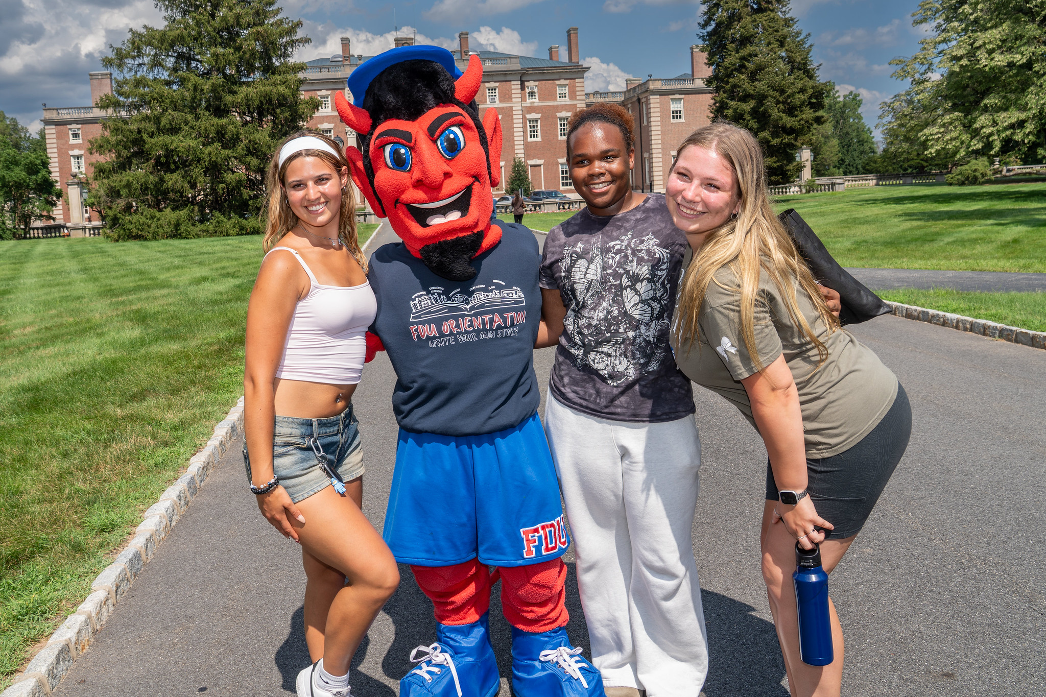 students and the FDU Ian the Devil mascot smile at the camera. the Hennessy Hall mansion stands in the background.