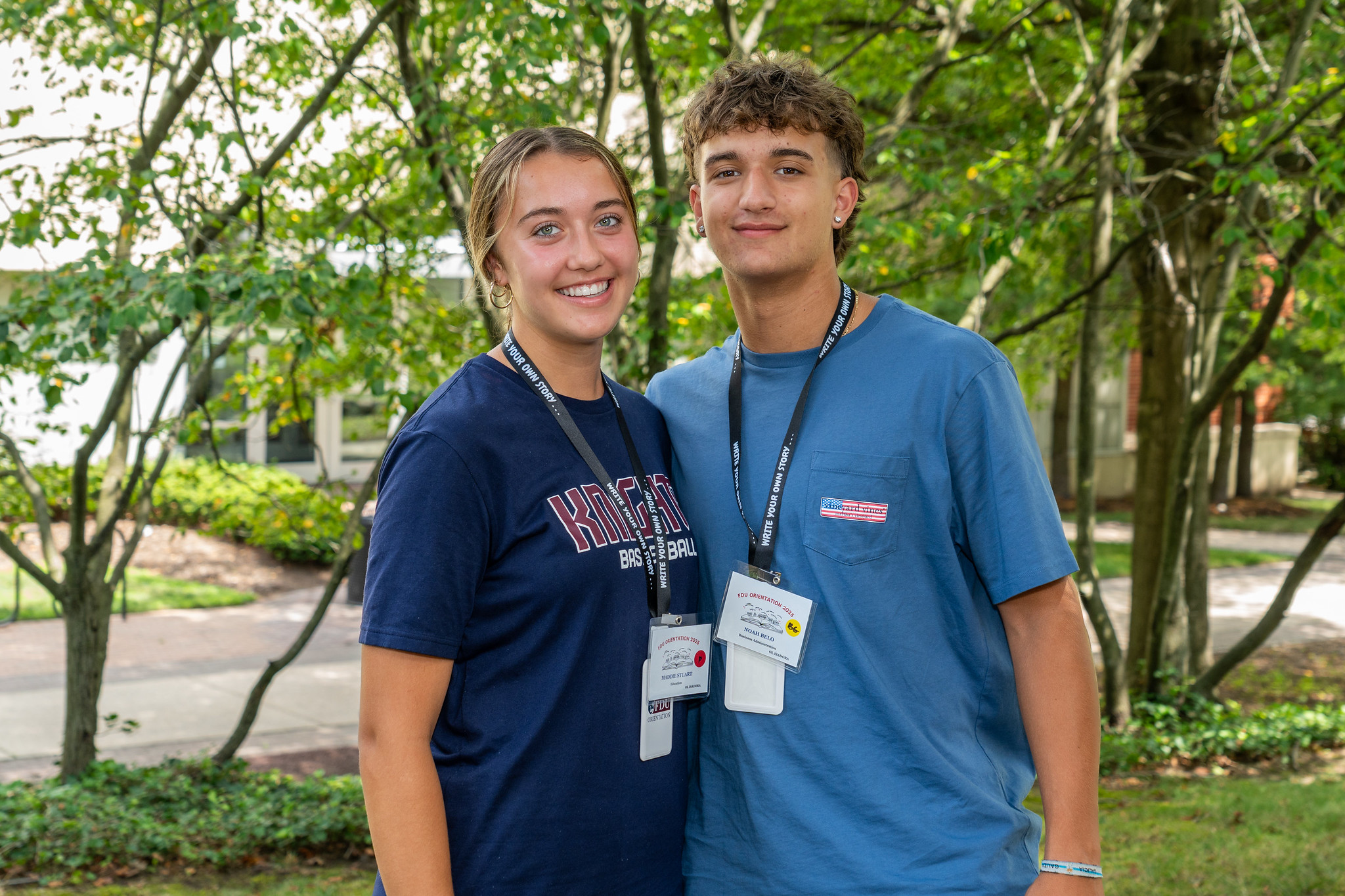 two students smile at the camera. they stand outside. one wears an FDU Knights Basketball shirt and they both wear orientation lanyards.