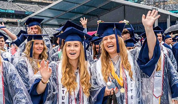 Two young women clad in caps and gowns smile and wave at graduation.
