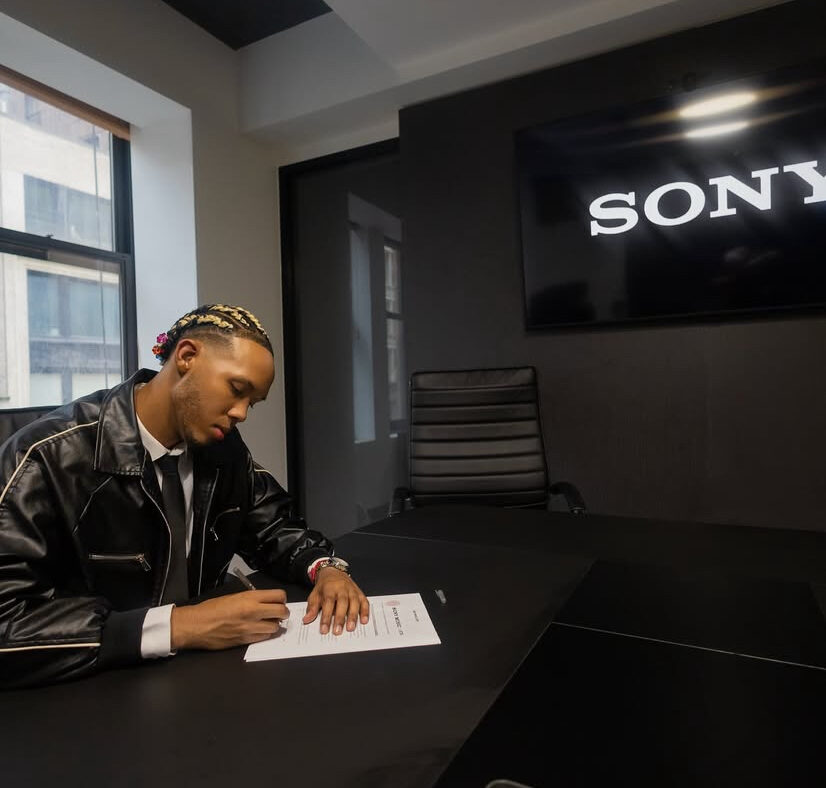 Claudio Balbi, also known artistically as Massivo, sits at a desk signing a contract with Sony Music/Latin. He is wearing a black leather jacket and tie, with braided hair. A large Sony logo is visible on a screen behind him.