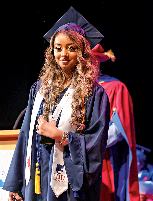 A young woman dressed in a graduation cap and gown stands on stage.