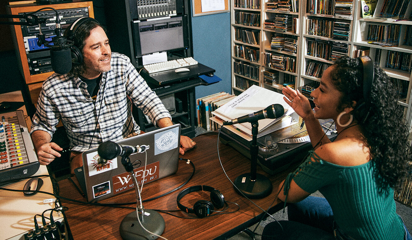 A man and a woman sit behind microphones in a radio station recording studio.