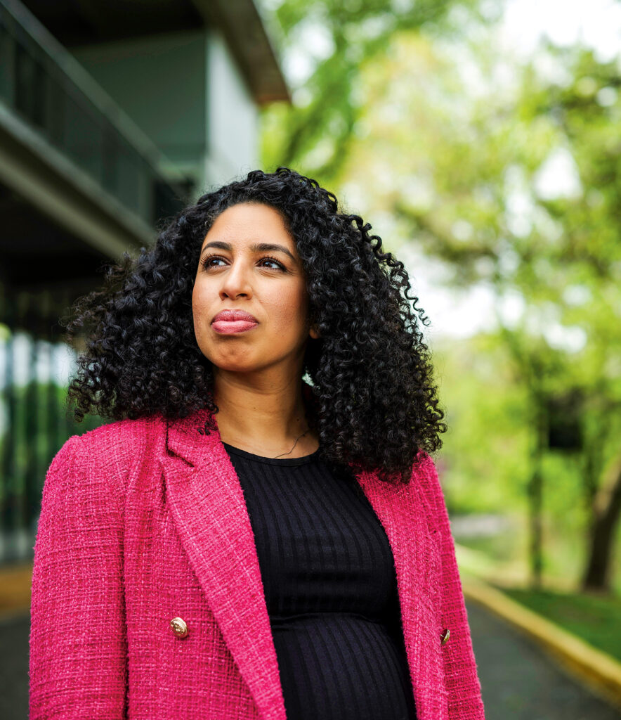 A woman wearing a pink blazer stands outside, gazing off into the distance.