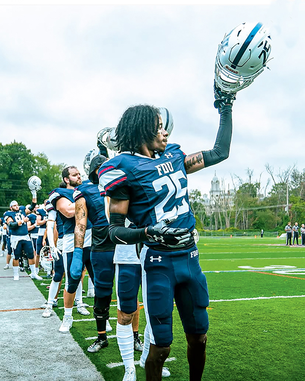 A line of young men in football uniforms hold up their helmets.