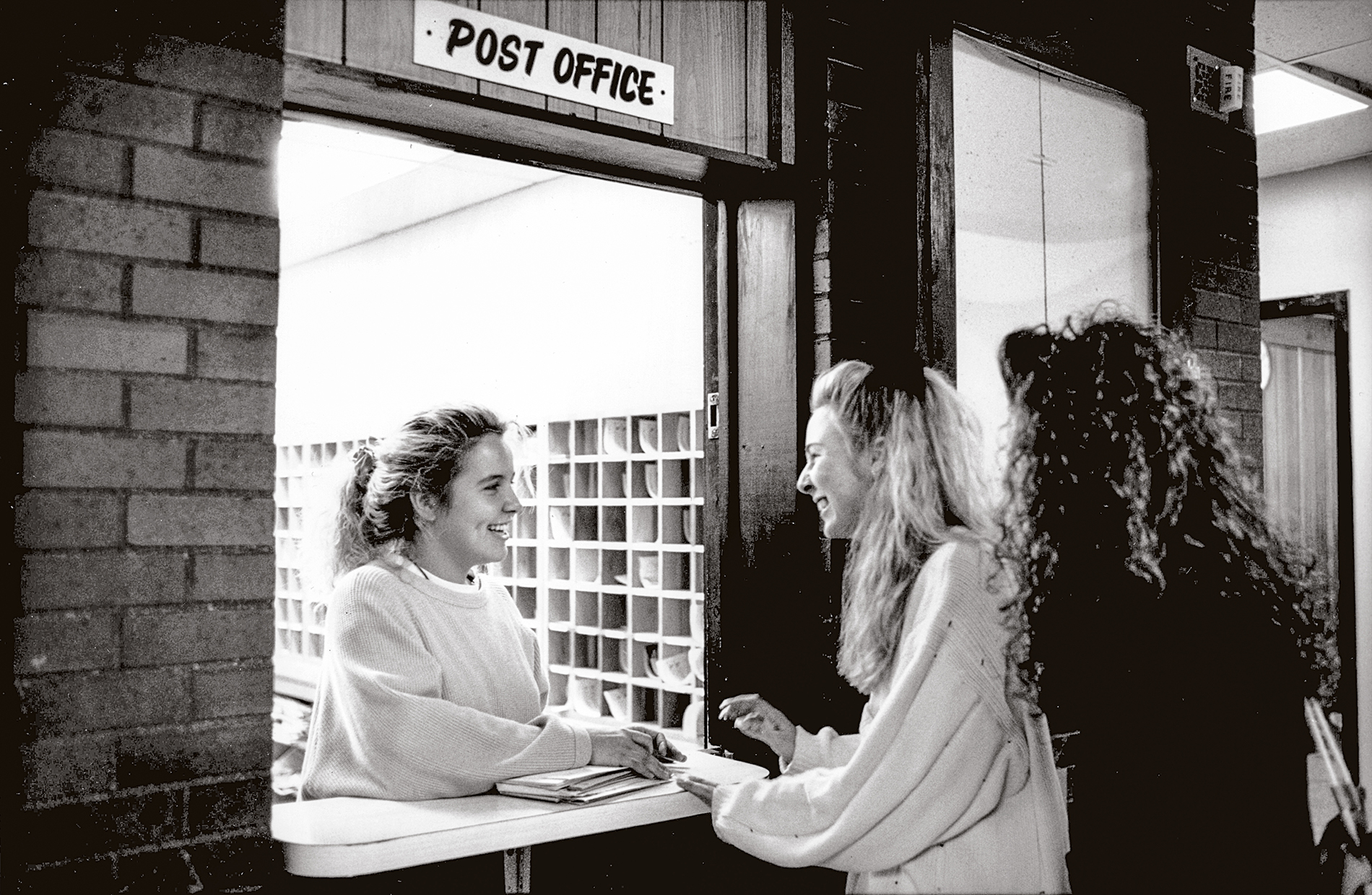 Two female college students pick up their mail at the campus post office from a woman in the mailroom.