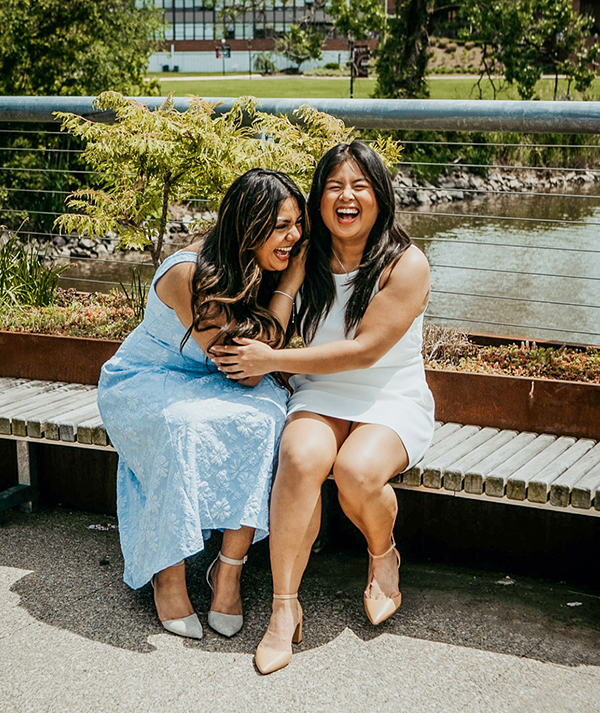 Two woman sit on a bench on a bridge over the river. They are hugging and laughing.