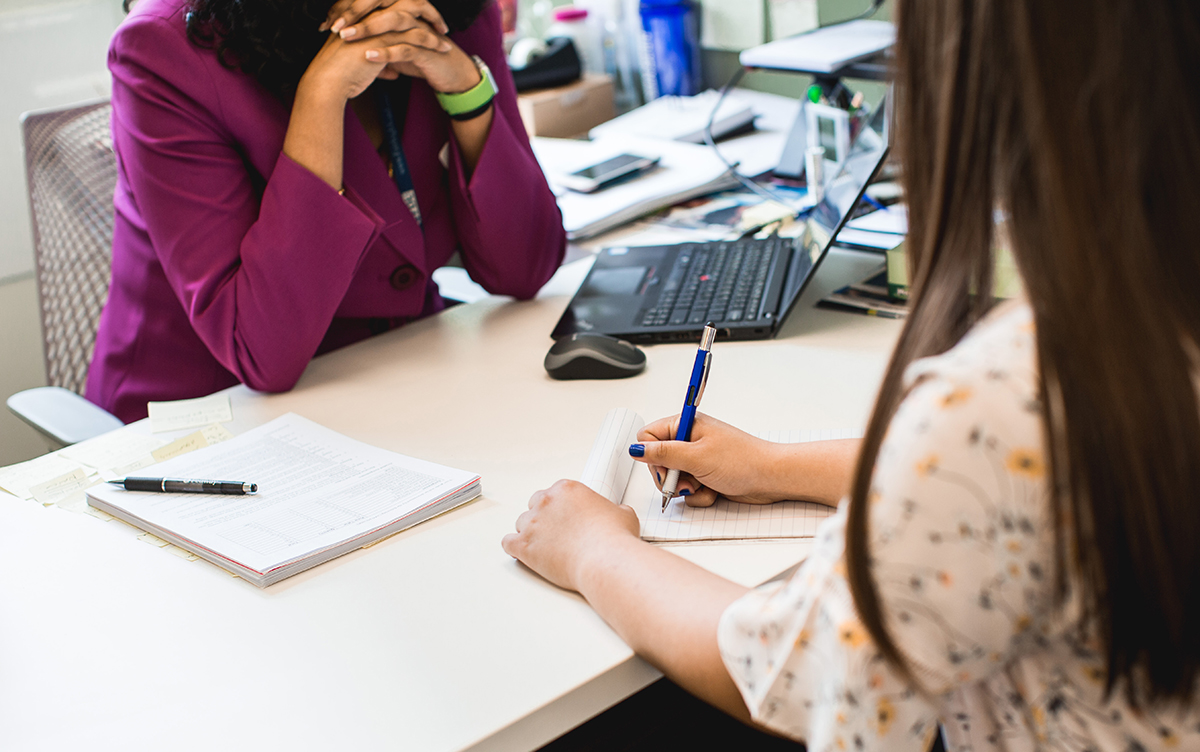 Two women sit at a desk in conference. One woman is taking notes.