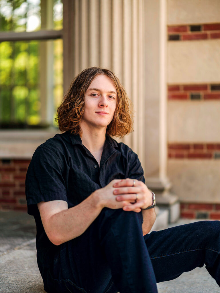 A young man sits on a concrete step outside and links his fingers together around one knee.
