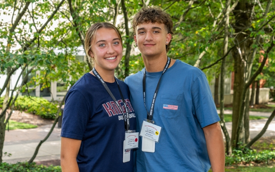 two students smile at the camera. they stand outside. one wears an FDU Knights Basketball shirt and they both wear orientation lanyards.