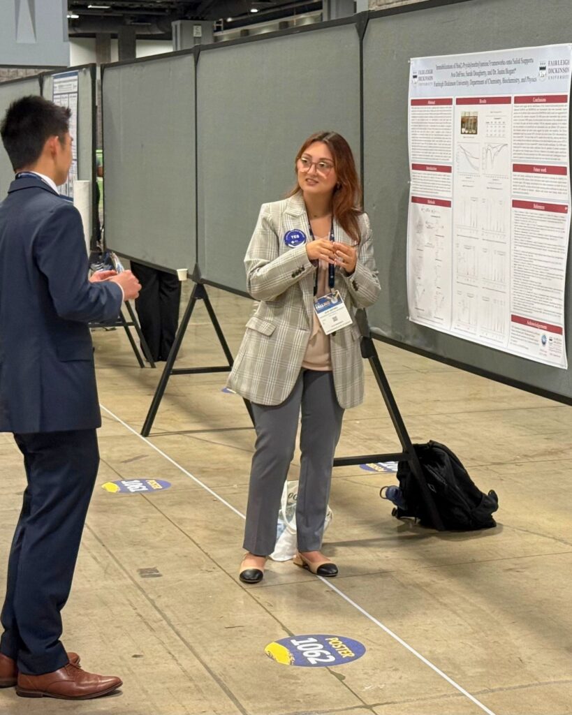 a college students presents to a colleague. the poster board presentation hangs behind her.