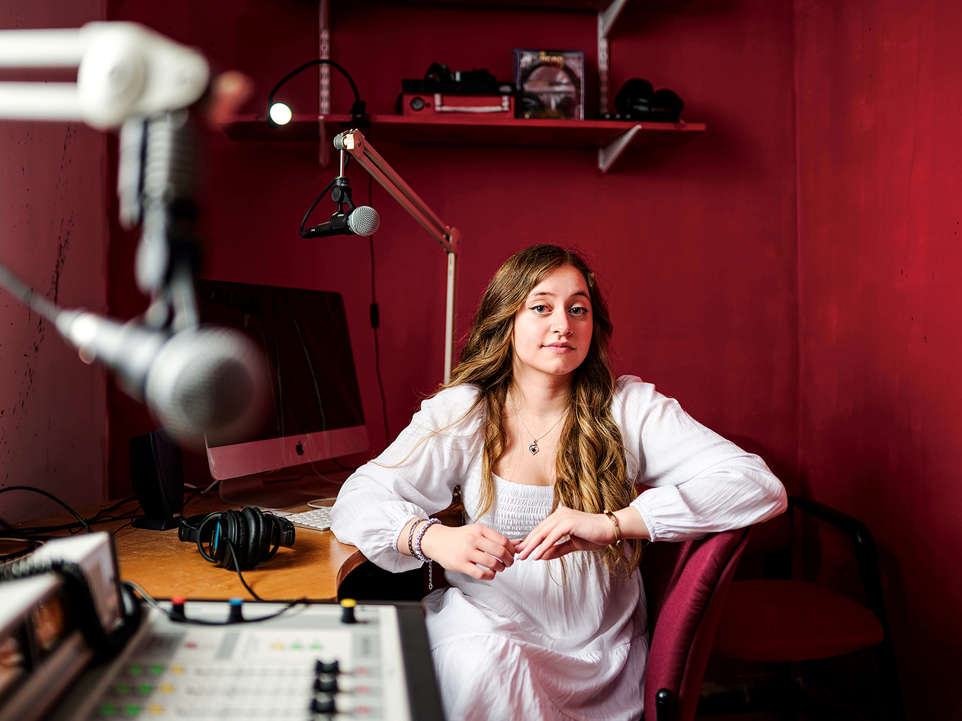 A young woman sits in a recording booth with podcast and radio equipment surrounding her.