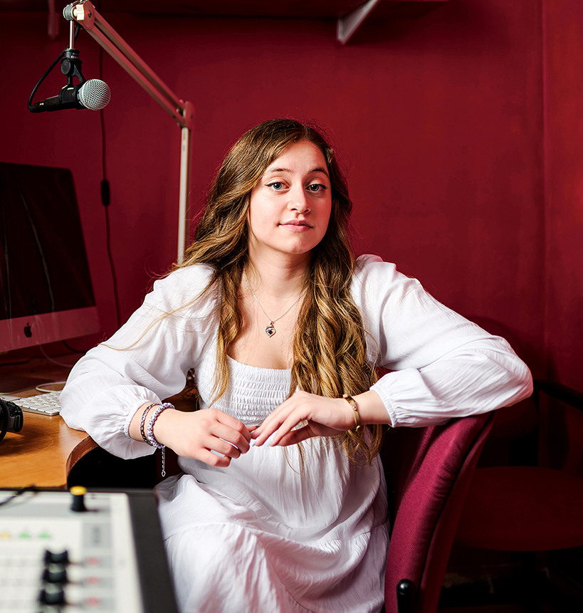 A young woman sits in a recording booth with podcast and radio equipment surrounding her.