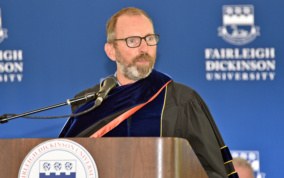 A man wearing academic regalia and glasses stands at a podium.
