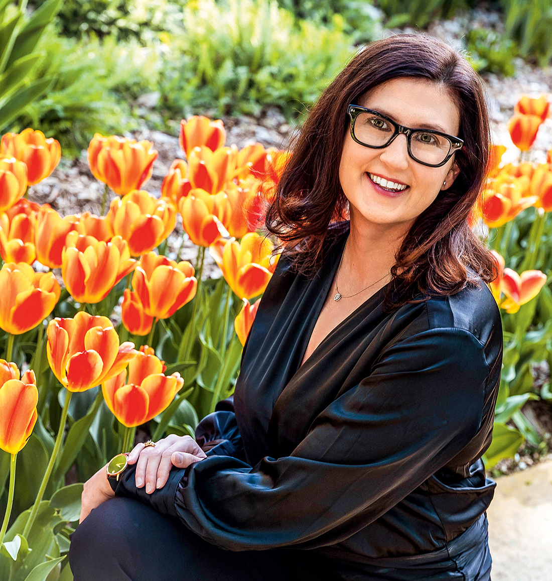 A woman wearing glasses sits in a garden near tulips.