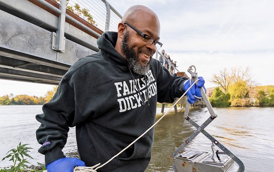 A man wearing an FDU sweatshirt and glasses uses research equipment in a river.
