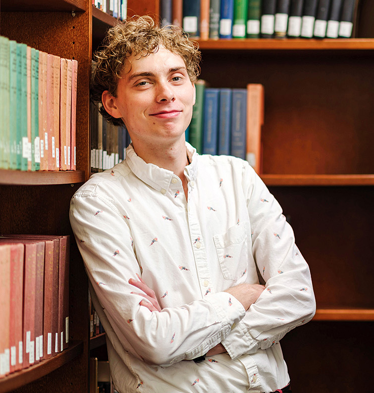 A young man crosses him arms and leans back against bookcases filled with texts.