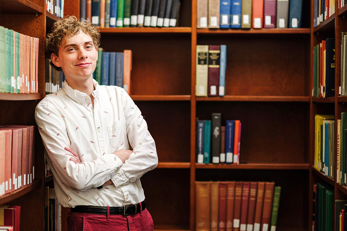 A young man crosses him arms and leans back against bookcases filled with texts.