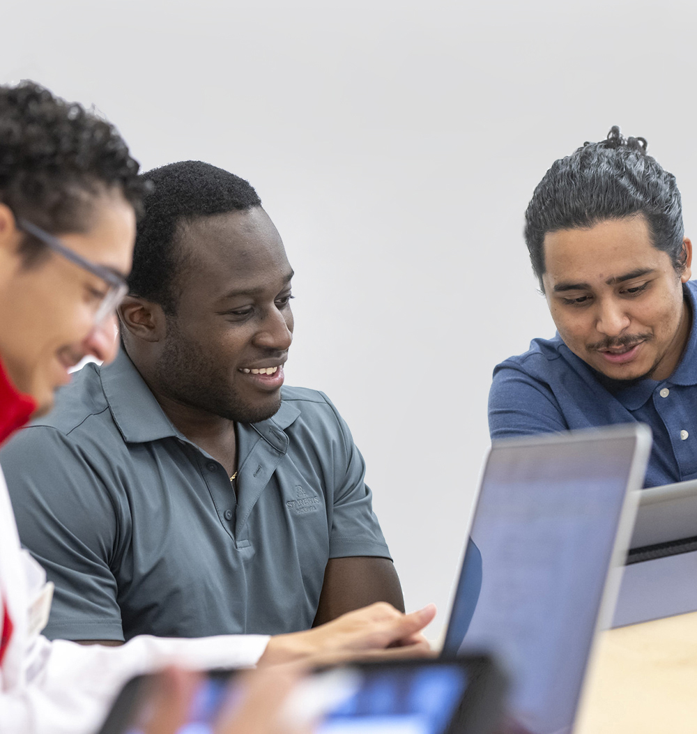 Three male students chat in class while working on their laptops.