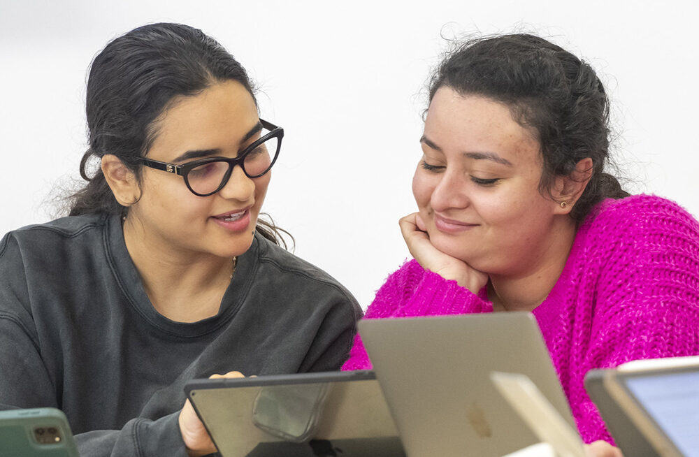 Two female students chat in class while working on their laptops.