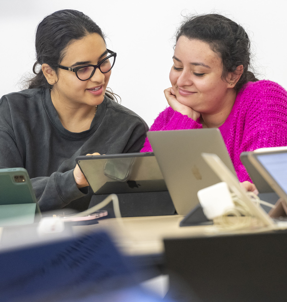 Two female students chat in class while working on their laptops.