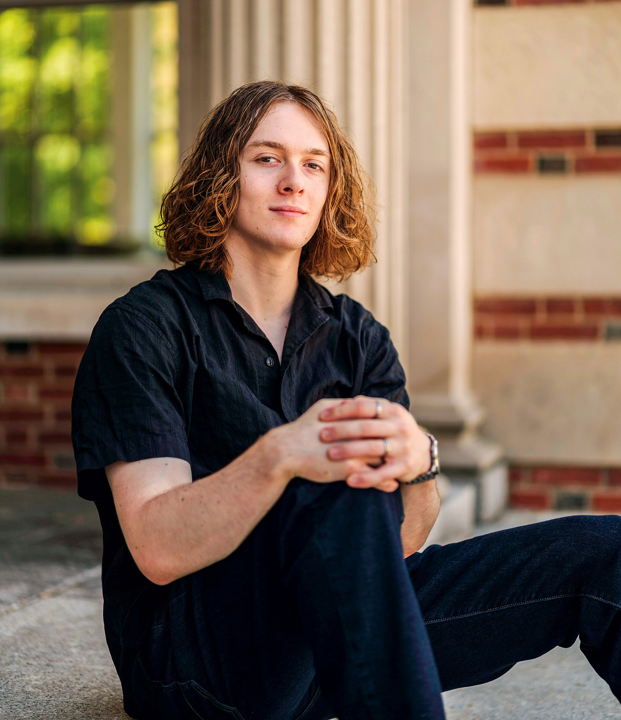 A young man sits on a concrete step outside and links his fingers together around one knee.