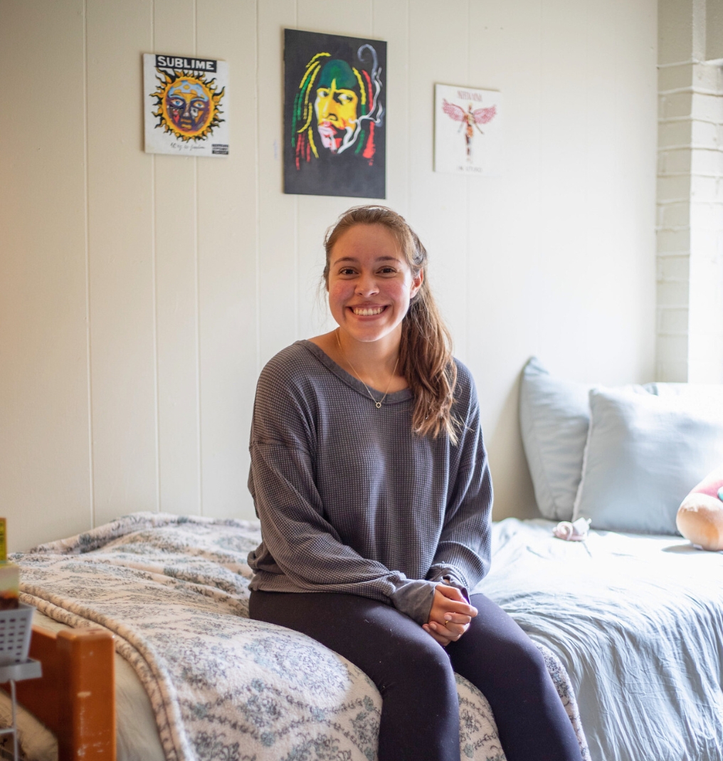 a college student sits on her dorm bed and smiles at the camera. there are music posters on the wall behind her.